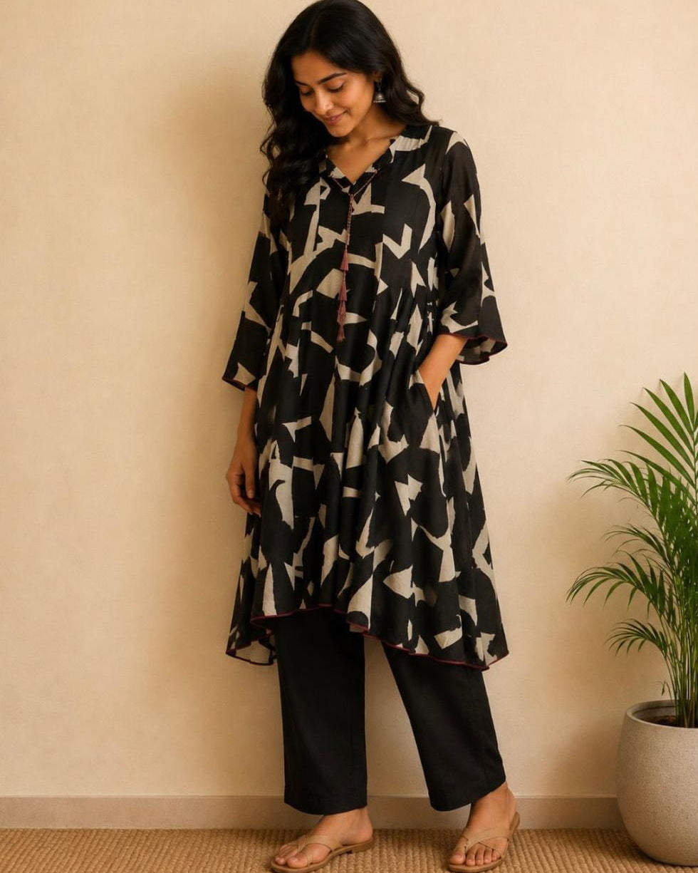 A woman wearing a black and beige abstract print kurta with black pants, standing in front of a beige wall next to a potted plant.