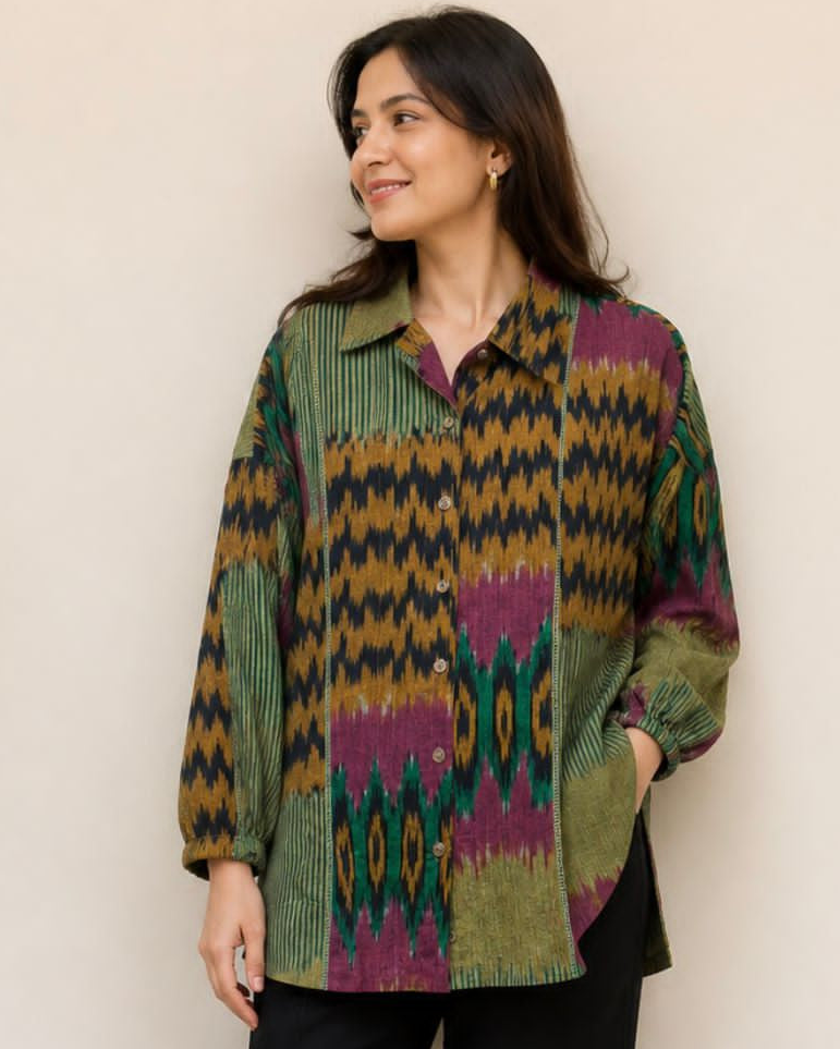 A woman modeling a long-sleeve button-up shirt with a colorful geometric ikat pattern in green, purple, yellow, and black, paired with black pants and sandals, standing against a plain beige wall.