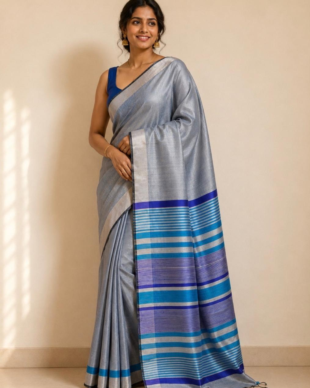 A woman modeling a light blue-gray silk saree with a blue and purple striped border, paired with a solid blue blouse, standing against a beige wall.
