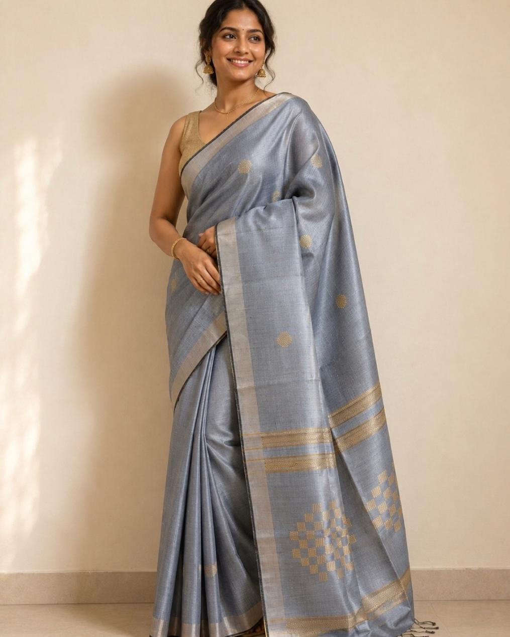 A woman modeling a light blue-gray silk saree with gold zari motifs and borders, paired with a matching sleeveless blouse, standing against a plain beige wall.