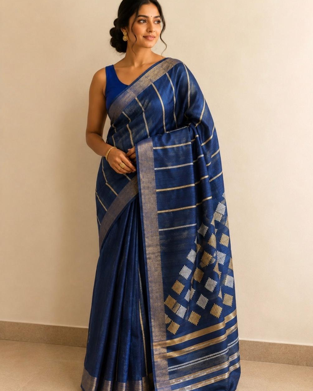 A woman modeling a royal blue silk saree with metallic gold and silver stripes and a geometric patterned border, paired with a matching blue blouse, set against a plain beige wall.