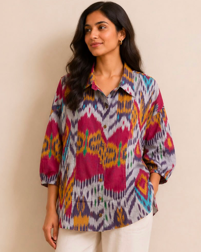 A woman modeling a colorful patterned button-up shirt with three-quarter sleeves, paired with white cropped pants and brown sandals, standing against a plain beige wall.