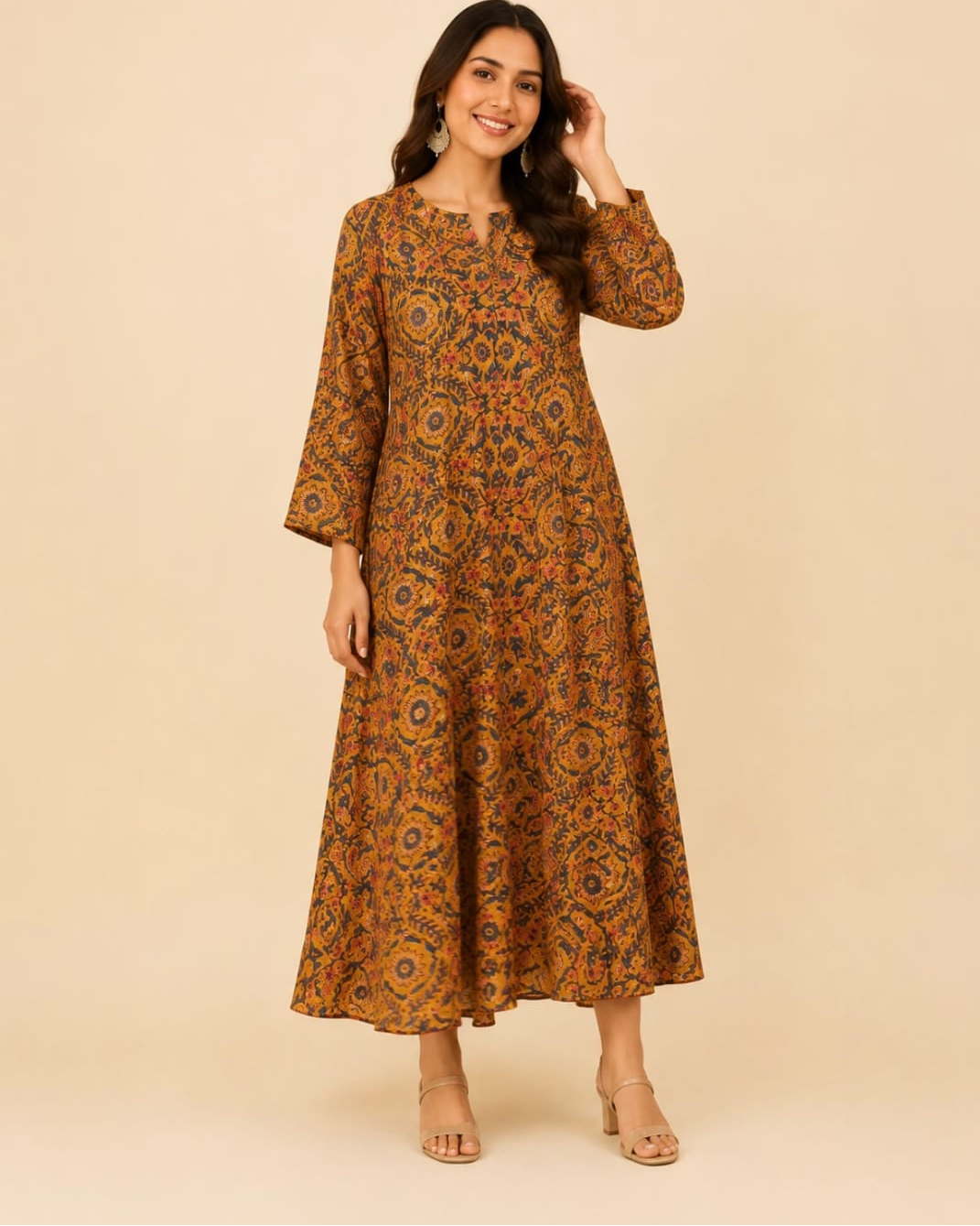 A woman modeling a long-sleeved, knee-length maxi dress with an intricate orange, brown, and blue floral mandala pattern against a solid beige studio background.