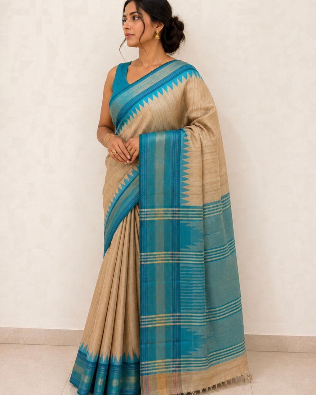 A woman modeling a beige and teal blue patterned cotton saree with a matching sleeveless blouse, standing against a plain off-white wall.