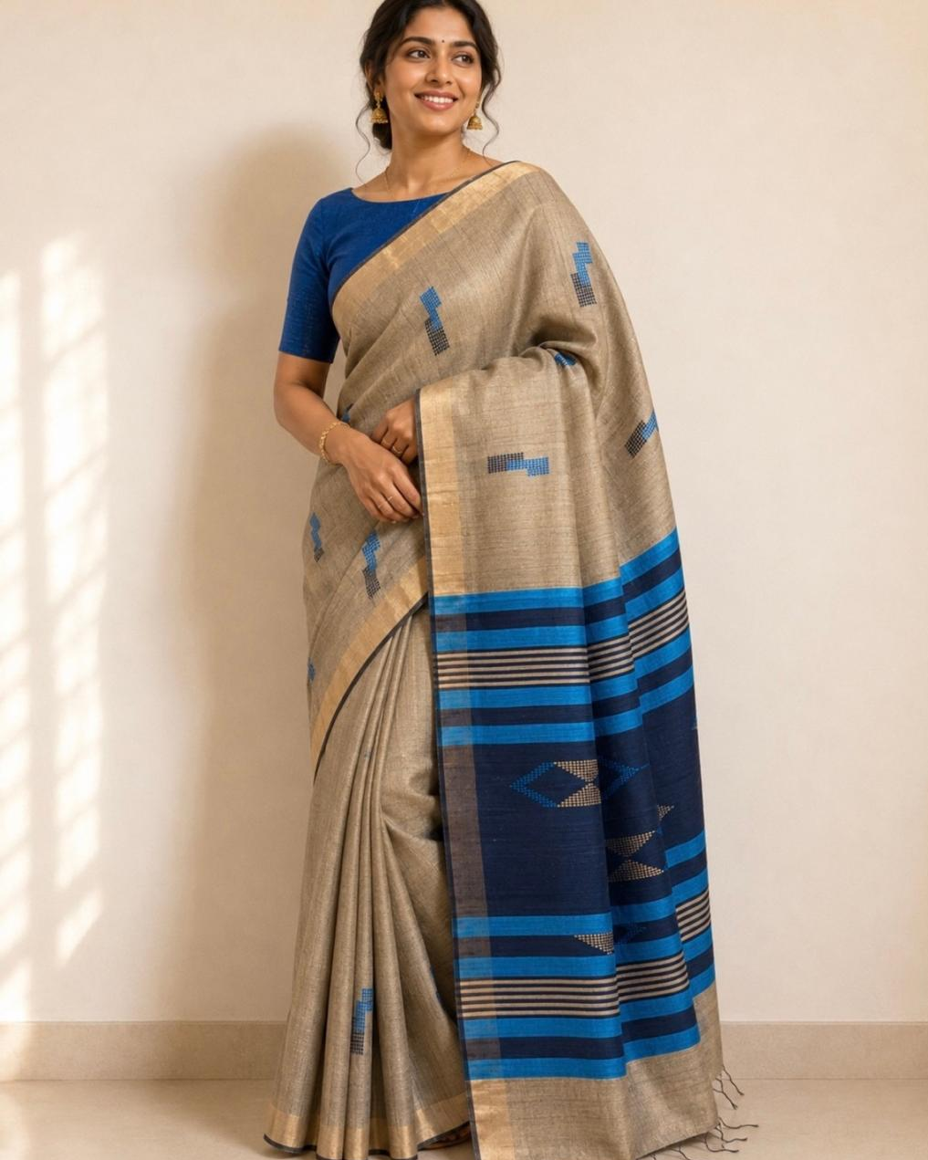 A woman modeling a beige and navy blue saree with geometric patterns, paired with a solid blue blouse, standing against a plain beige wall.