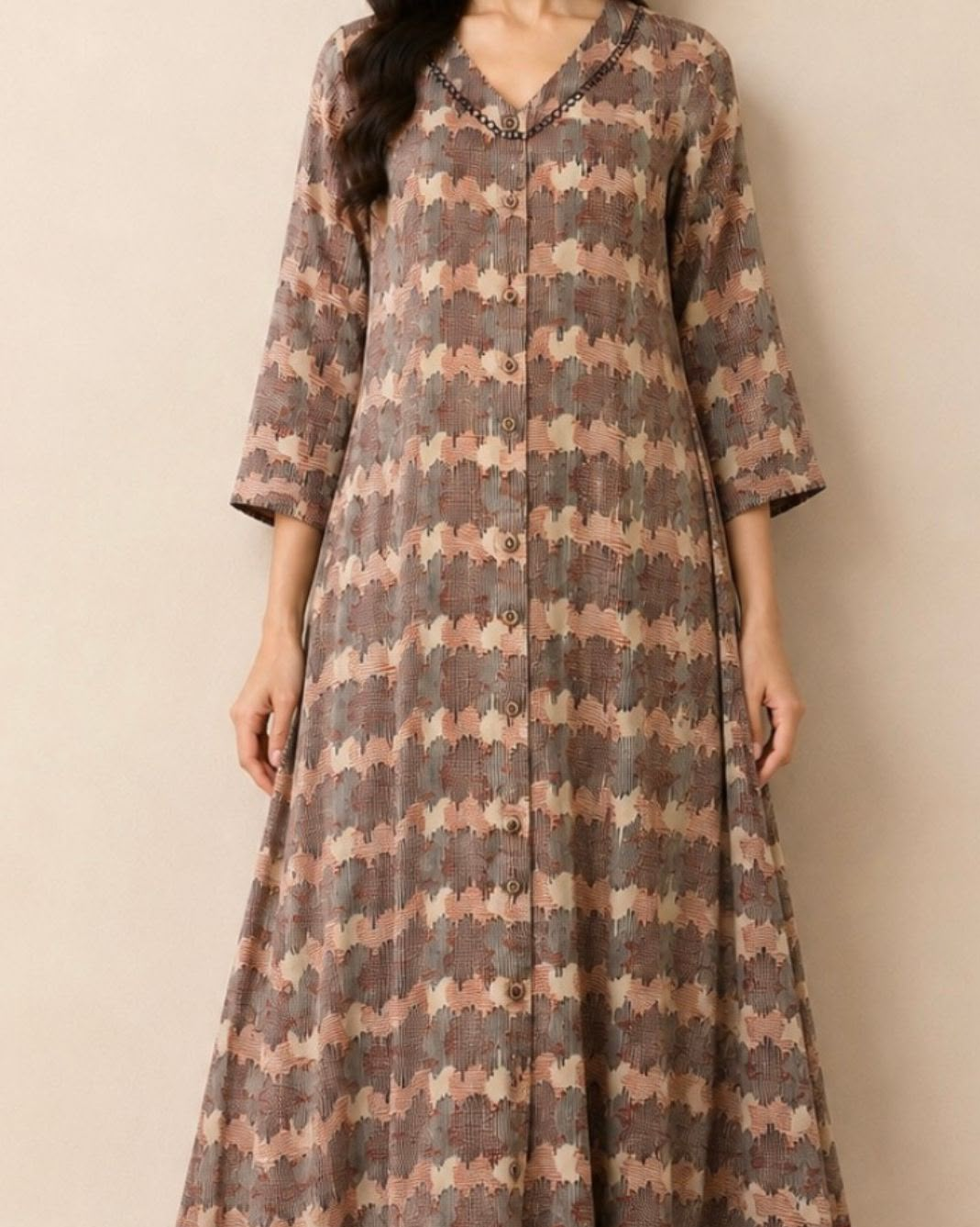 A woman wearing a long-sleeved, button-front kaftan dress with a brown, beige, and pink geometric pattern, shown against a plain beige background.