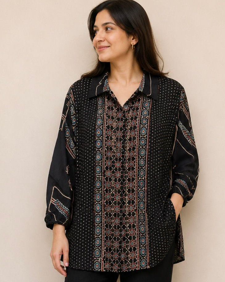 A woman modeling a black long-sleeve button-up shirt with intricate brown and beige geometric patterns, paired with black pants and sandals, standing against a plain beige wall.