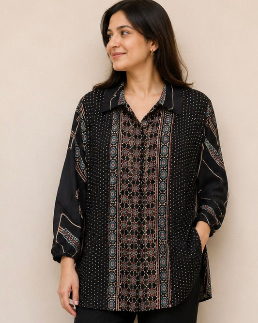 A woman modeling a black long-sleeve button-up shirt with intricate brown and beige geometric patterns, paired with black pants and sandals, standing against a plain beige wall.