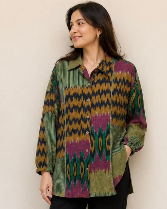 A woman modeling a long-sleeve button-up shirt with a colorful geometric ikat pattern in green, purple, yellow, and black, paired with black pants and sandals, standing against a plain beige wall.