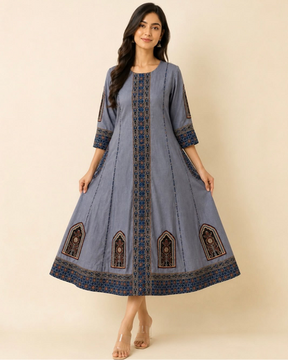 A woman modeling a dusty blue A-line kurta with intricate dark blue and brown geometric embroidery on the front panel, cuffs, and hem, set against a solid beige studio background.