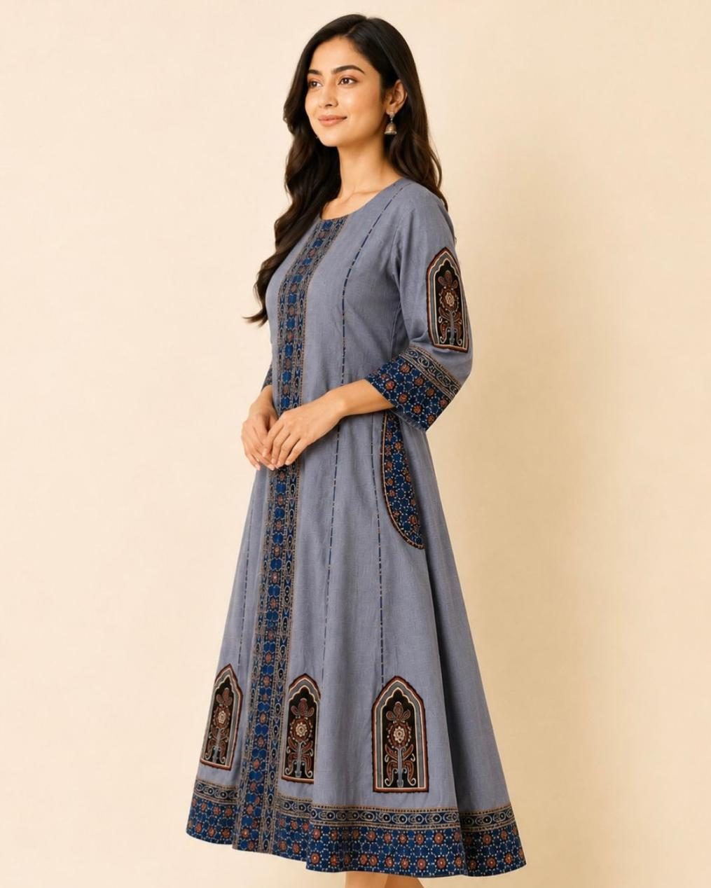 A woman modeling a dusty blue A-line kurta with intricate blue, brown, and gold geometric embroidery on the front, sleeves, and hem, set against a solid beige background.