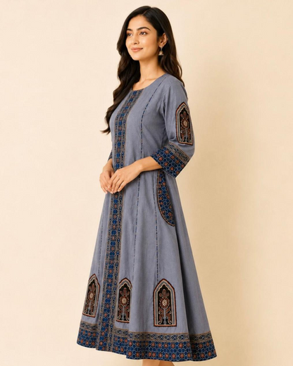 A woman modeling a dusty blue A-line kurta with intricate blue, brown, and gold geometric embroidery on the front, sleeves, and hem, set against a solid beige background.