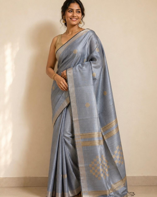 A woman modeling a light blue-gray silk saree with gold zari motifs and borders, paired with a matching sleeveless blouse, standing against a plain beige wall.