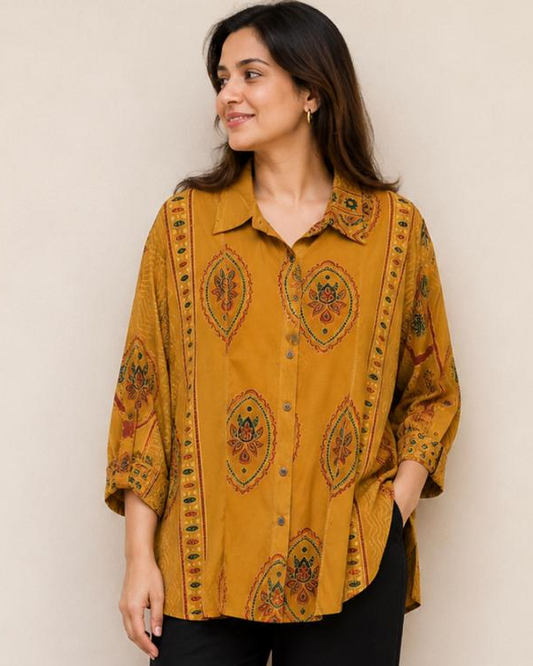 A woman wearing a mustard yellow button-up shirt with a brown and red paisley pattern, paired with black pants and brown sandals, standing against a plain beige wall.