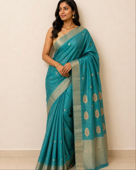 A woman modeling peacock blue-green silk saree with a beige border and gold motifs, standing against a plain beige wall.
