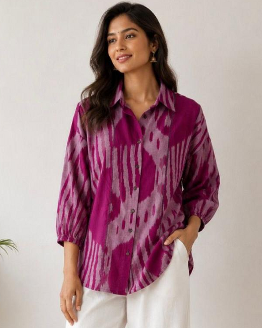 A woman modeling a purple and pink ikat patterned button-down shirt with white wide-leg pants, standing in a minimalist room with a potted plant on the left.