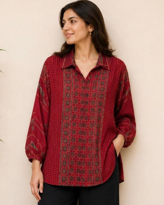 A woman stands indoors wearing a long-sleeve collared blouse with a red and black embroidered pattern and black wide-leg pants, paired with tan sandals. A potted plant is visible at the left edge against a plain beige wall.
