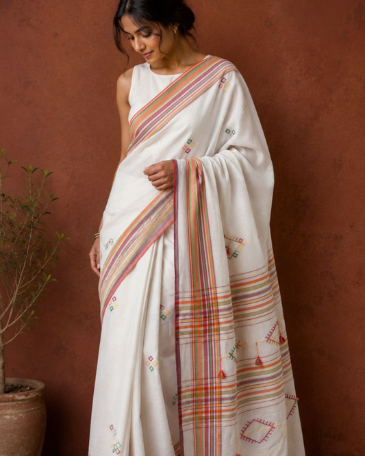 A woman wearing a white silk saree with colorful striped borders and small embroidered motifs, standing against a textured terracotta wall.