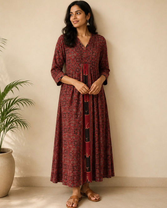 A woman wearing a long-sleeved, V-neck maxi dress with a red and black floral pattern, standing against a beige wall next to a potted plant.